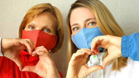 Old and young woman standing side by side with masks on and creating heart shapes with their hands