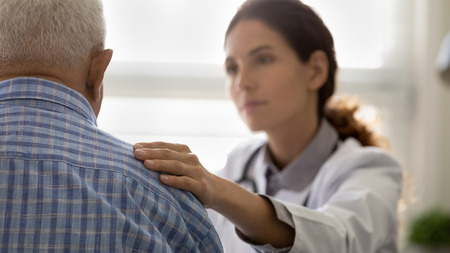 Compassionate female Doctor talking to patient who is an elderly man, her hand is on his shoulder